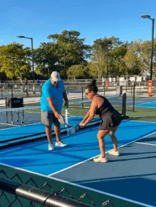 Person in blue shirt teaches another person pickleball on an outdoor court under clear skies.