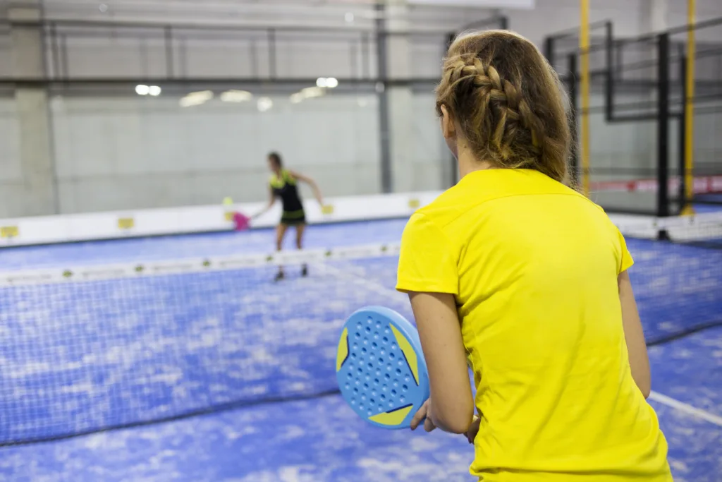 Two women playing padel on a blue court, one in yellow holding a racket, the other blurred in the distance.