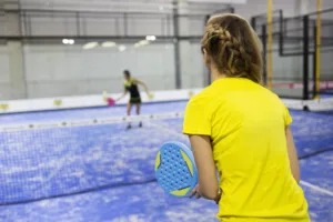 Two women playing padel on a blue court, one in yellow holding a racket, the other blurred in the distance.