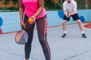 Two people playing pickleball on a court, one holding a paddle and an orange ball.